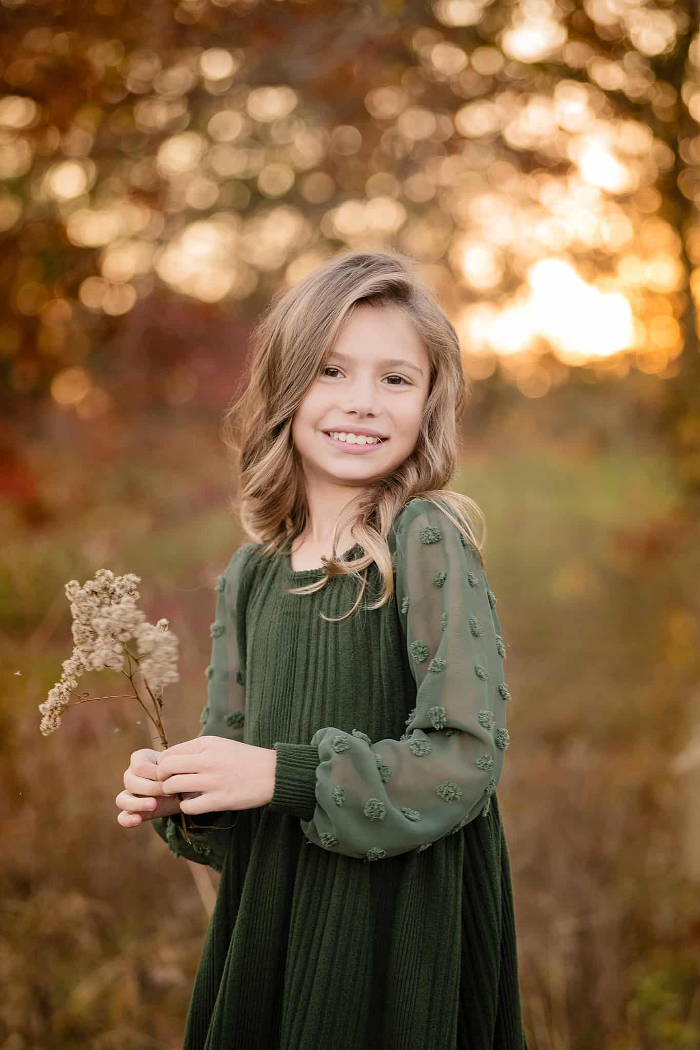 A smiling young girl in a green dress playing with wildflowers in a park at sunset